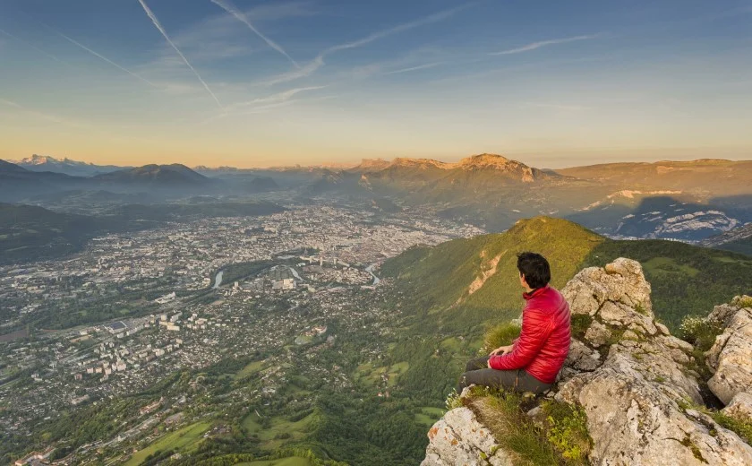 Un homme en doudoune rouge est assis en haut du Saint-Eynard et regarde l'agglomération grenobloise en bas.