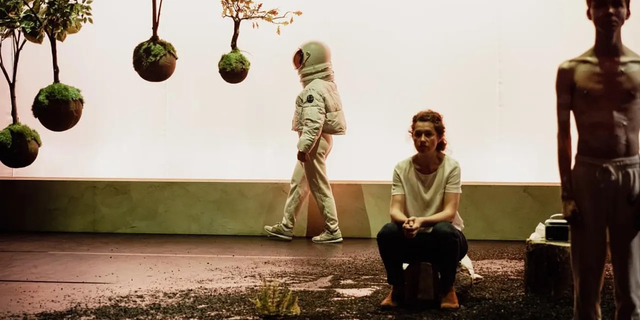 Photo couleur où on voit sur la gauche des plantes suspendues et sur la droite un astronaute, une femme et un jeune homme un