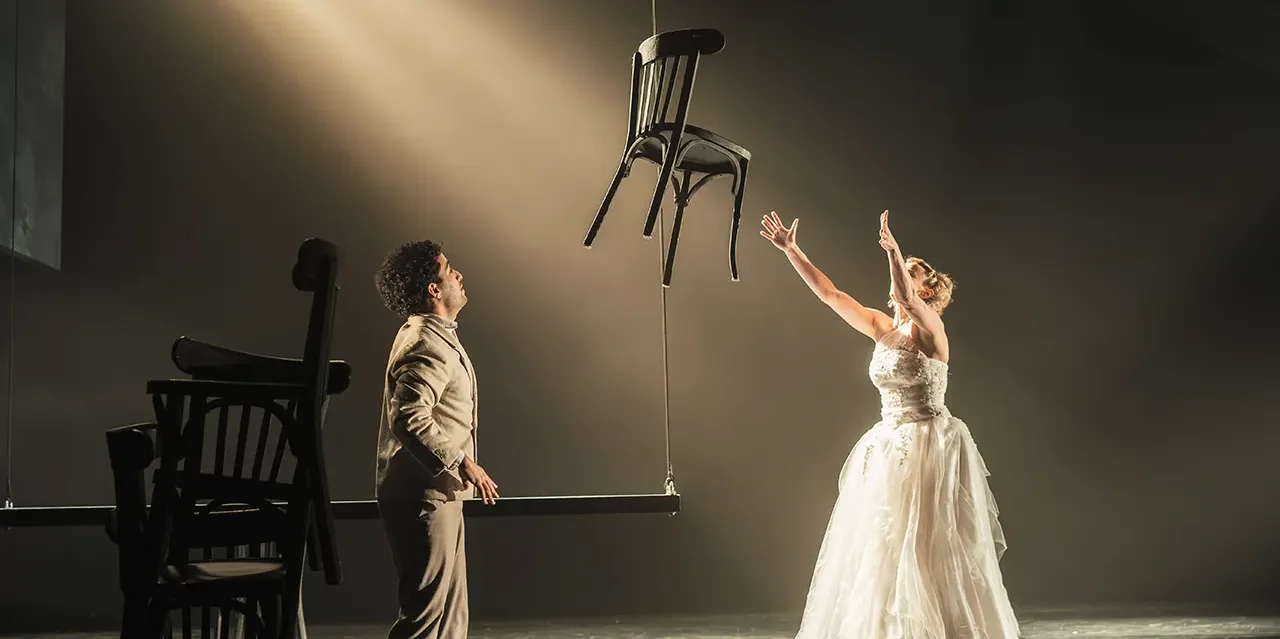 Scène du spectacle "Ombres portées". Un homme et une femme en robe de mariée regardent une chaise qui flotte dans les airs